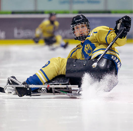 Eine Person in gelber Eishockeyausr stung sitzt auf einem Schlitten und f hrt auf dem Eis, w hrend im Hintergrund eine weitere Person zu sehen ist. Beschreibung von KI generiert