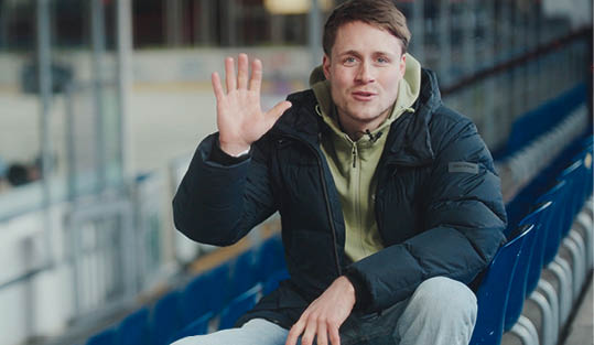 Eine Person in einer schwarzen Jacke und einem hellgr nen Kapuzenpullover sitzt auf blauen Sitzreihen und hebt die Hand zum Gru . Beschreibung von KI generiert