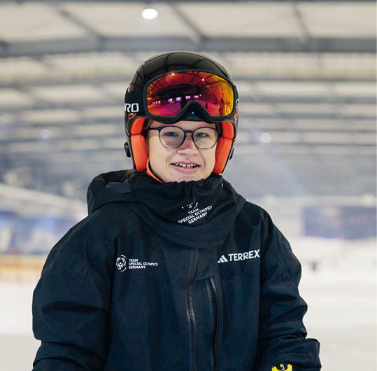 Person tr gt Skihelm, Skibrille und eine schwarze Jacke mit Logos der Special Olympics Germany und Adidas Terrex in einer Indoor-Skihalle. Beschreibung von KI generiert