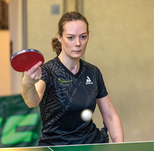 Eine Person in einem schwarzen Sportshirt spielt Tischtennis und schl gt den Ball mit einem roten Schl ger. Beschreibung von KI generiert