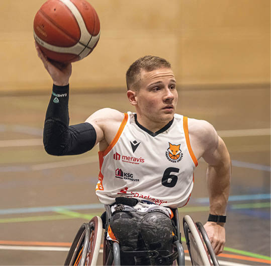 Eine Person in einem Sportrollstuhl tr gt ein wei es Basketballtrikot mit der Nummer sechs und h lt einen Basketball in der erhobenen Hand in einer Sporthalle. Beschreibung von KI generiert