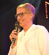 Eine Person mit kurzen Haaren und Brille spricht in ein Mikrofon und tr gt eine helle, flauschige Jacke vor dunklem Hintergrund. Beschreibung von KI generiert