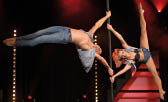 Zwei Erwachsene in l ssiger Kleidung f hren eine akrobatische Pole-Dance-Performance auf einer B hne mit roter Beleuchtung und schwarzen Hintergrund durch. Beschreibung von KI generiert
